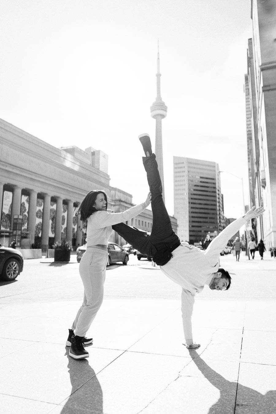 A playful moment captured in black and white, featuring a man performing a handstand with the help of a woman, set against the backdrop of the CN Tower in Toronto.