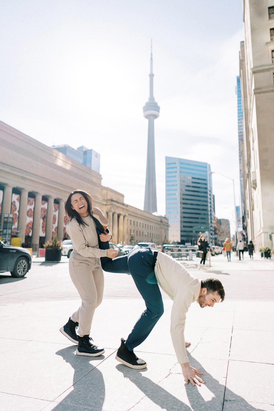 Two people share a playful moment in front of the CN Tower in Toronto, with one bending over and the other laughing joyfully.