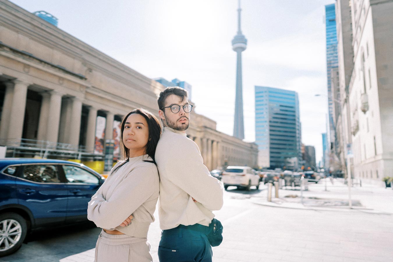 A man and woman pose back to back on a Toronto street, with the CN Tower visible in the background.
