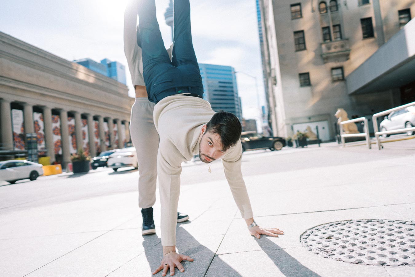 A man performs a handstand on a city street, supported by another person, with urban buildings in the background.