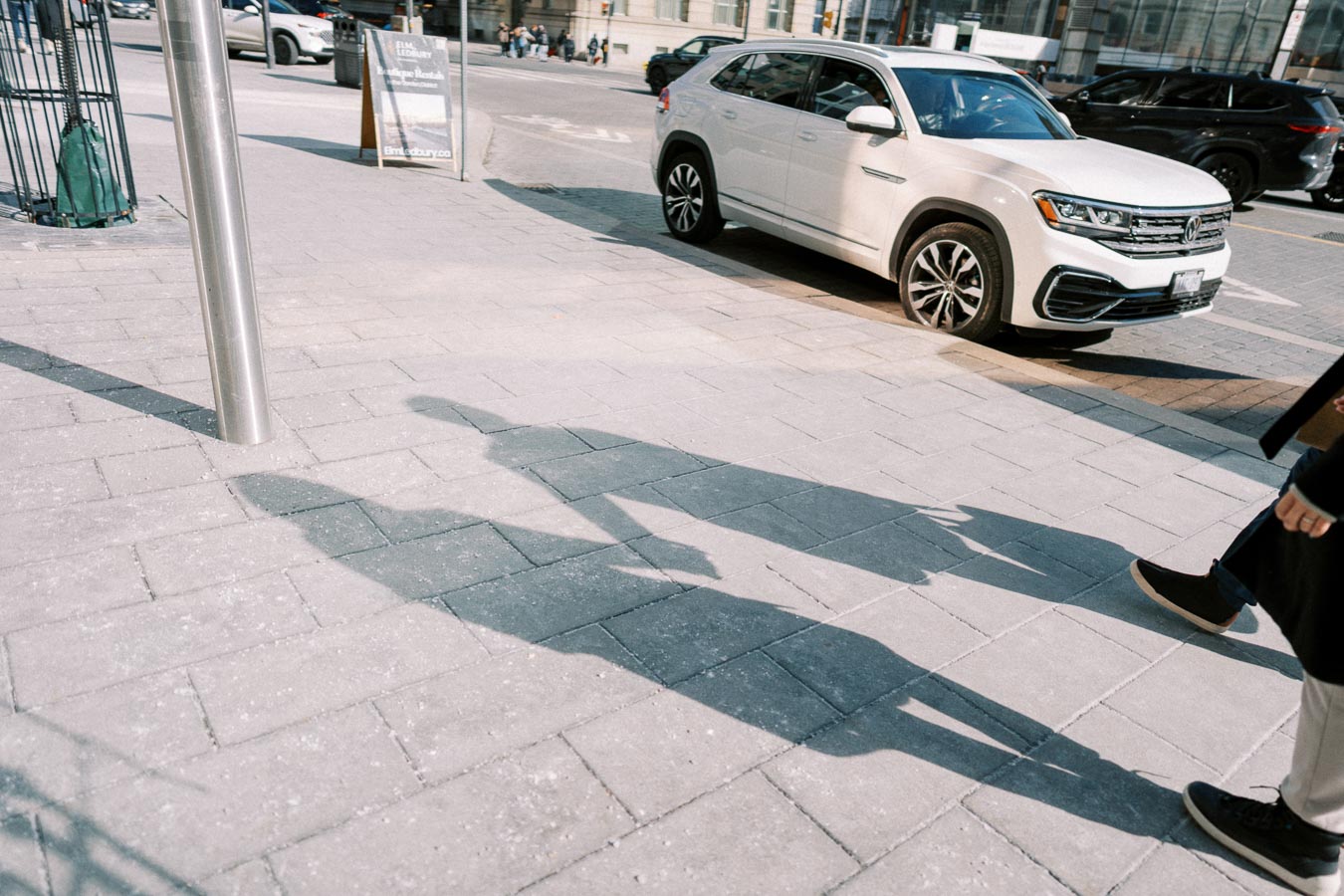 A white Volkswagen SUV is parked on a city street, while the shadows of two people walking are cast on the sidewalk.
