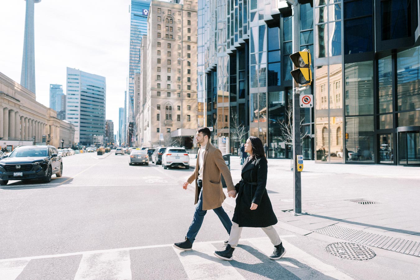 A couple crosses a street in downtown Toronto, with the CN Tower visible in the background.