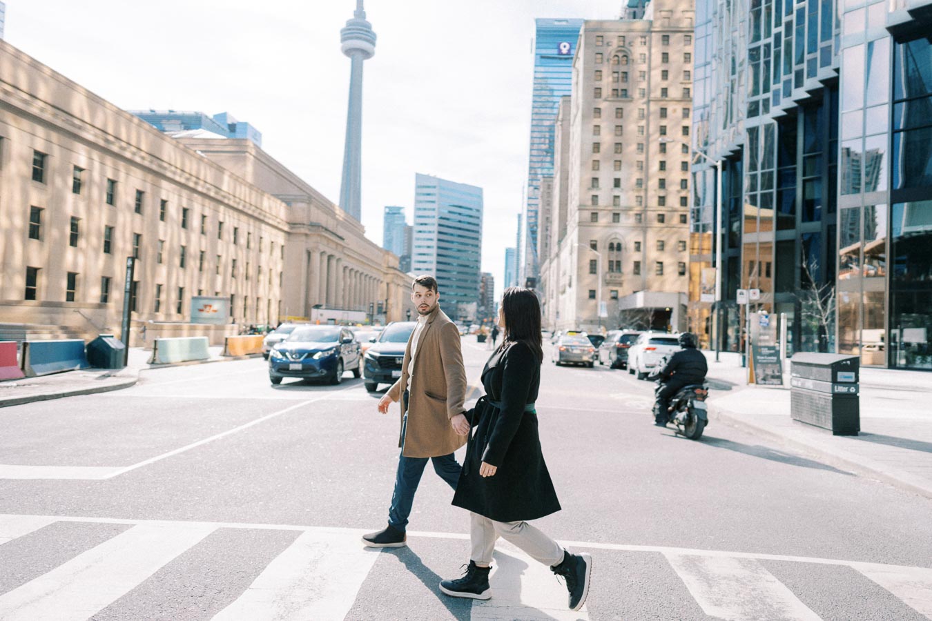 A couple walks across a street in Toronto, with the iconic CN Tower visible in the background, surrounded by urban architecture and traffic.