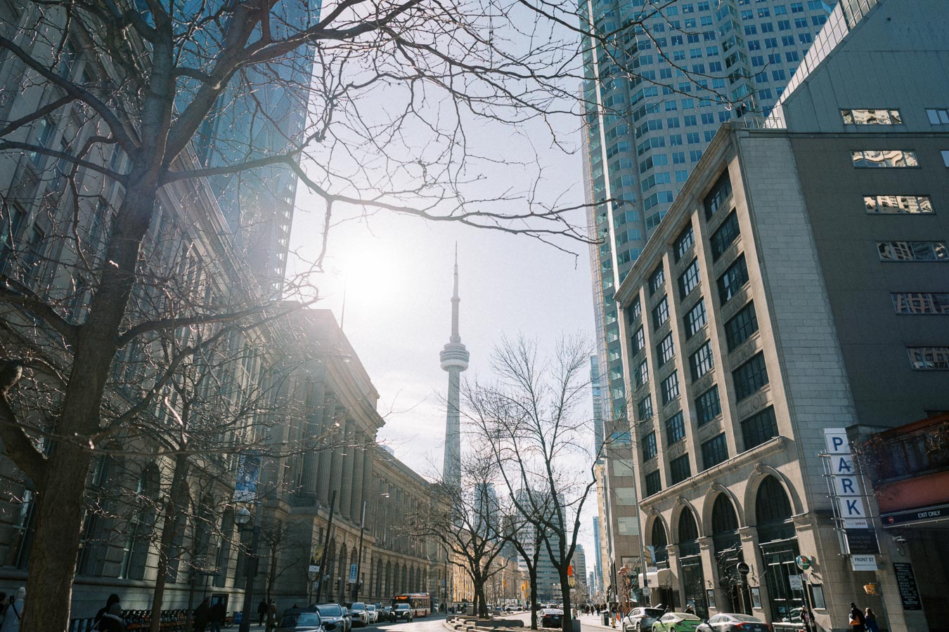 A street view in Toronto featuring the iconic CN Tower in the background, surrounded by tall buildings and bare trees, under a clear sky.