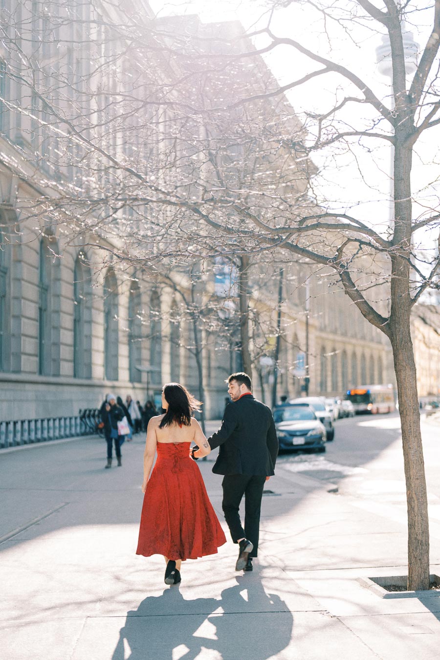 A couple strolls down a sunlit city street, with the woman wearing a red dress and the man in a black suit, framed by urban architecture and bare trees.