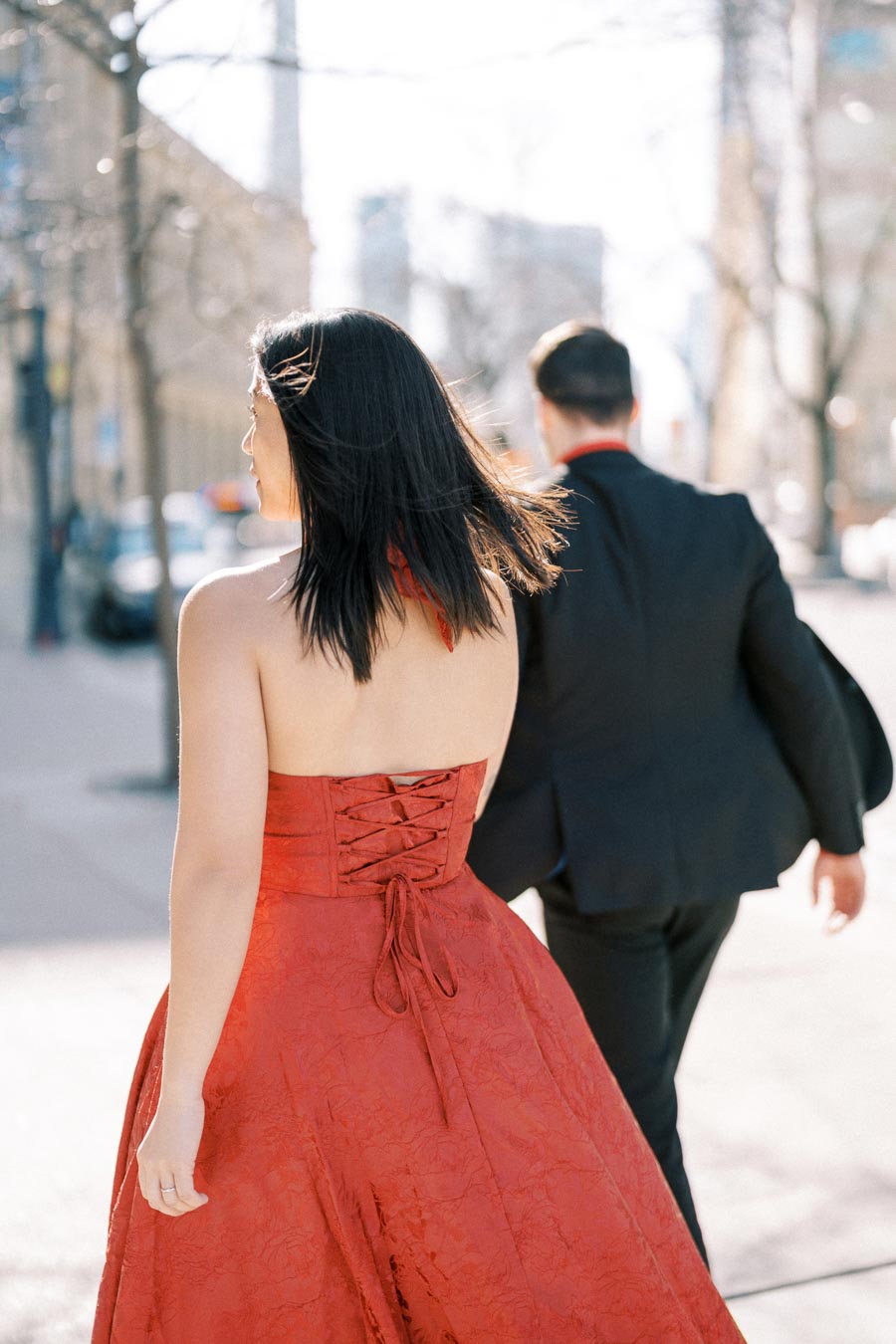 A woman in a red dress and a man in a black suit walk together on a sunny city street.