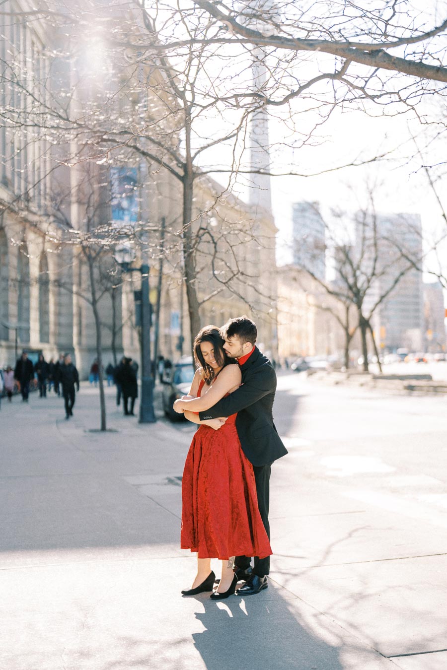 A couple embraces on a sunlit city street, with the woman wearing a red dress and the man in a black suit, surrounded by urban architecture and bare trees.