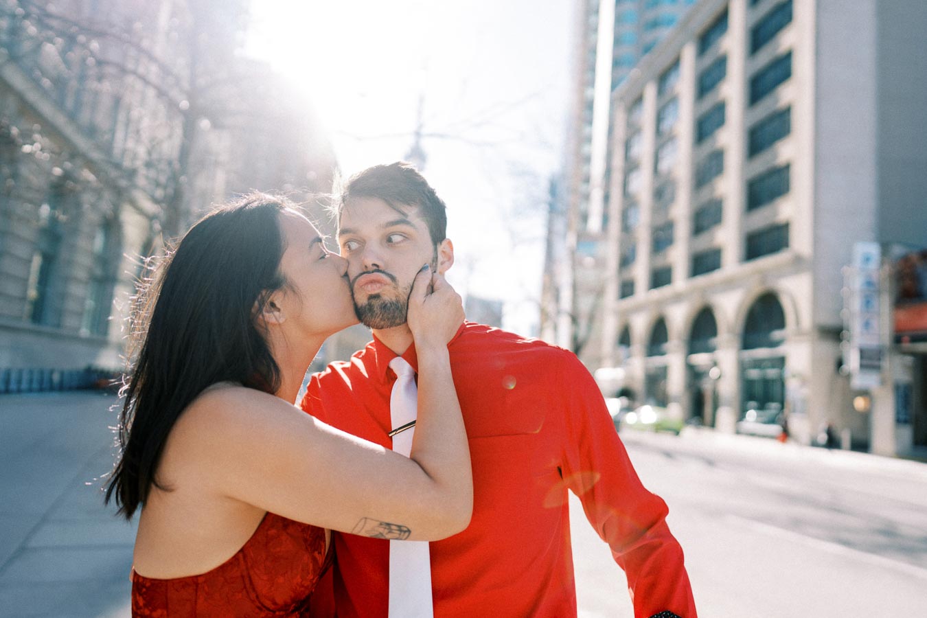 A woman playfully kisses a man on the cheek in a city street, with sunlight streaming in the background. The man is wearing a red shirt and white tie, while the woman is dressed in a red outfit.