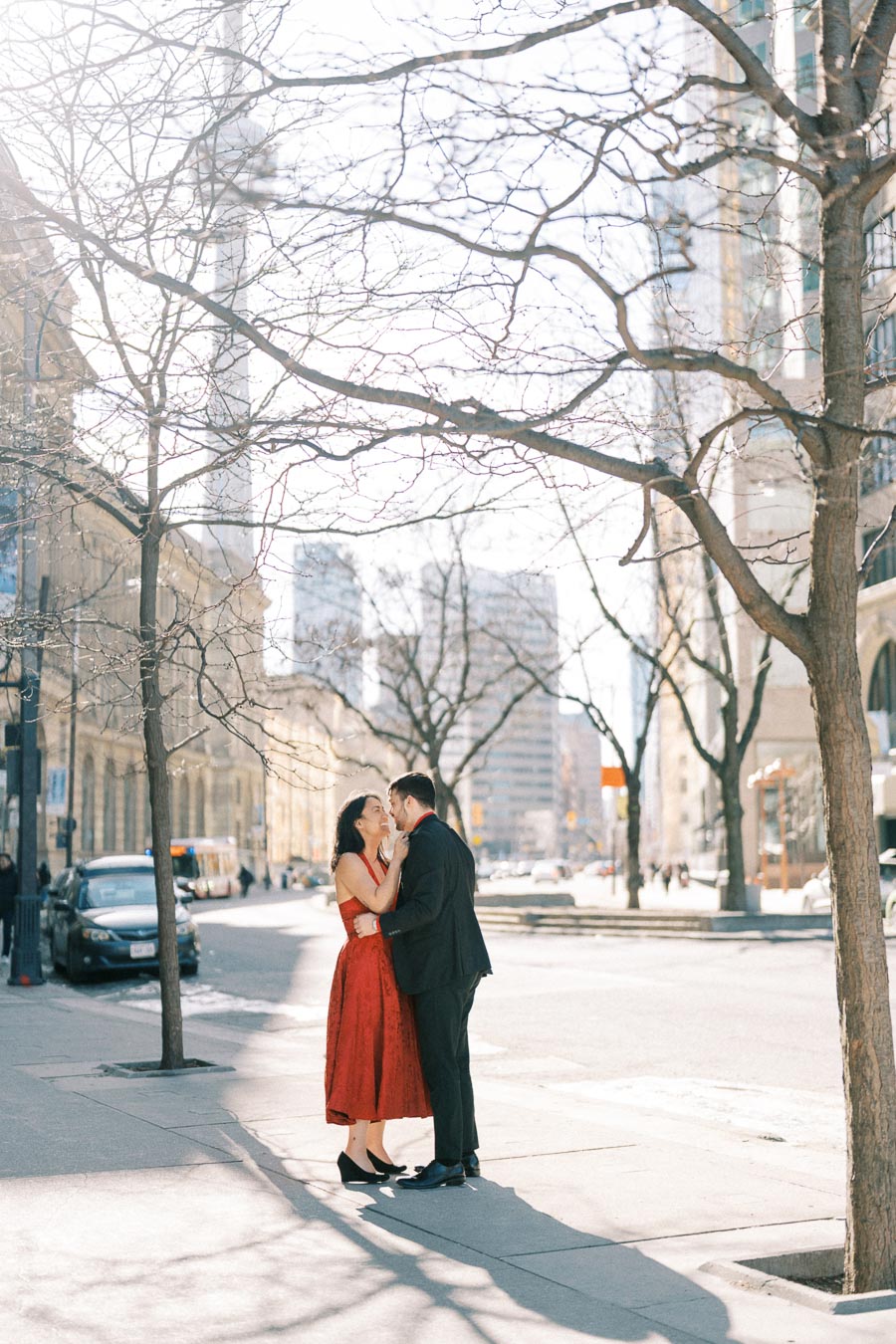 A couple shares a tender moment on a city street, surrounded by bare trees and urban architecture.