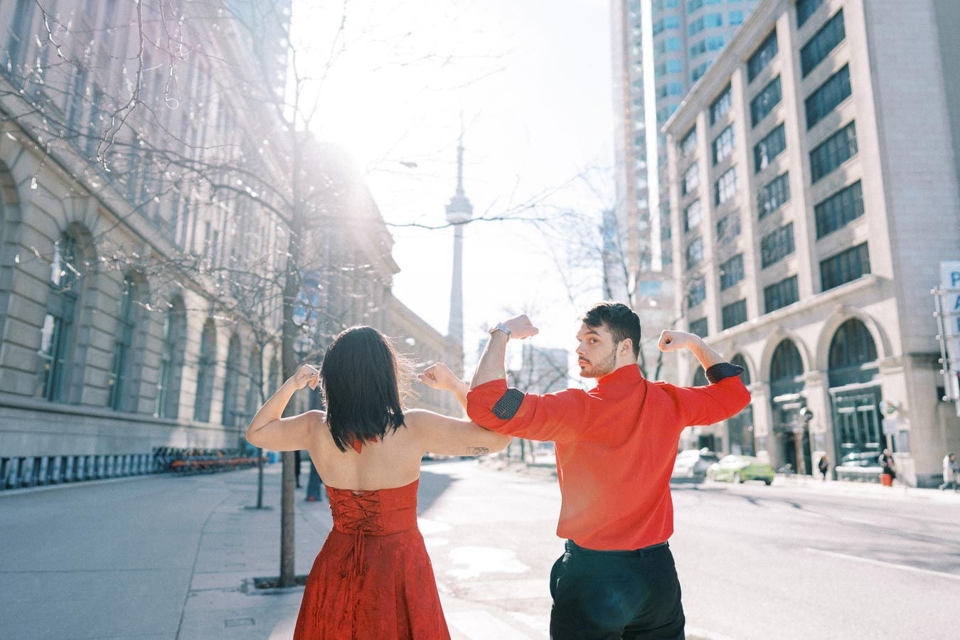 A couple in red clothing strikes a playful pose on a city street with the CN Tower in the background, capturing a sunny day in Toronto.