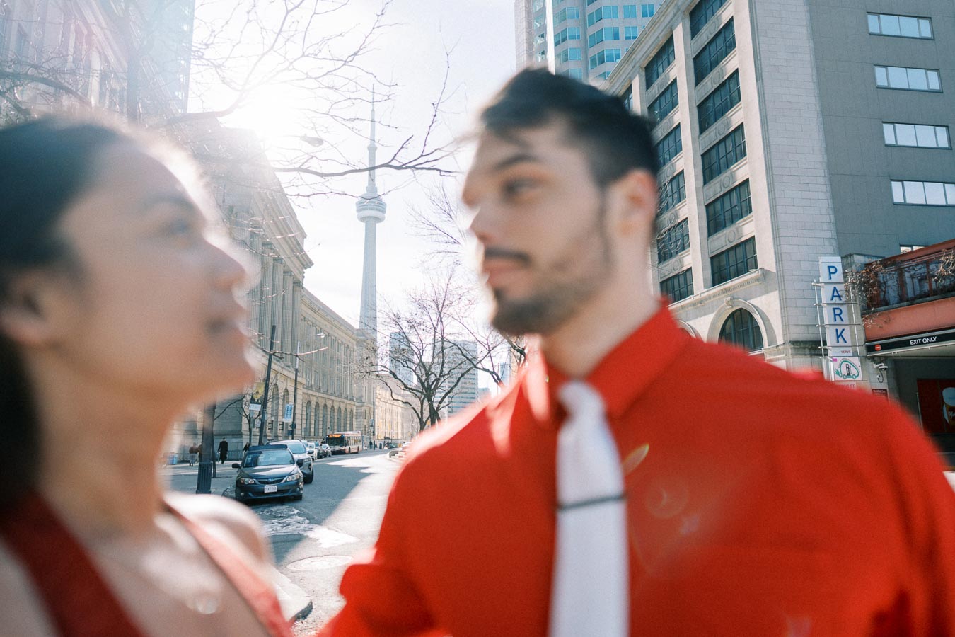 A couple in red attire stands on a city street with the CN Tower visible in the background, capturing a sunny day in Toronto.