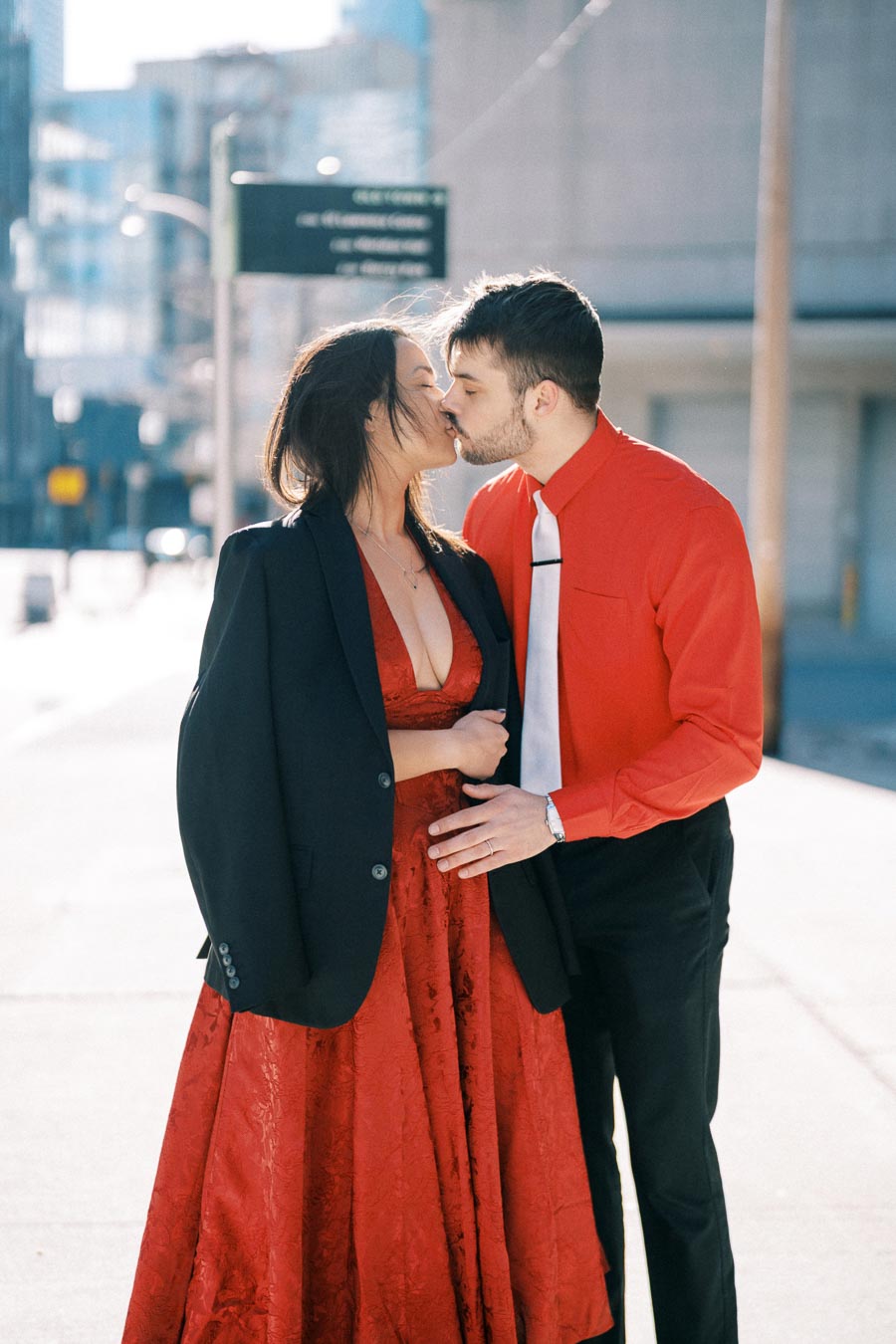 A couple shares a kiss on a city street, dressed in matching red attire. The woman wears a red dress with a black blazer, while the man sports a red shirt with a white tie.