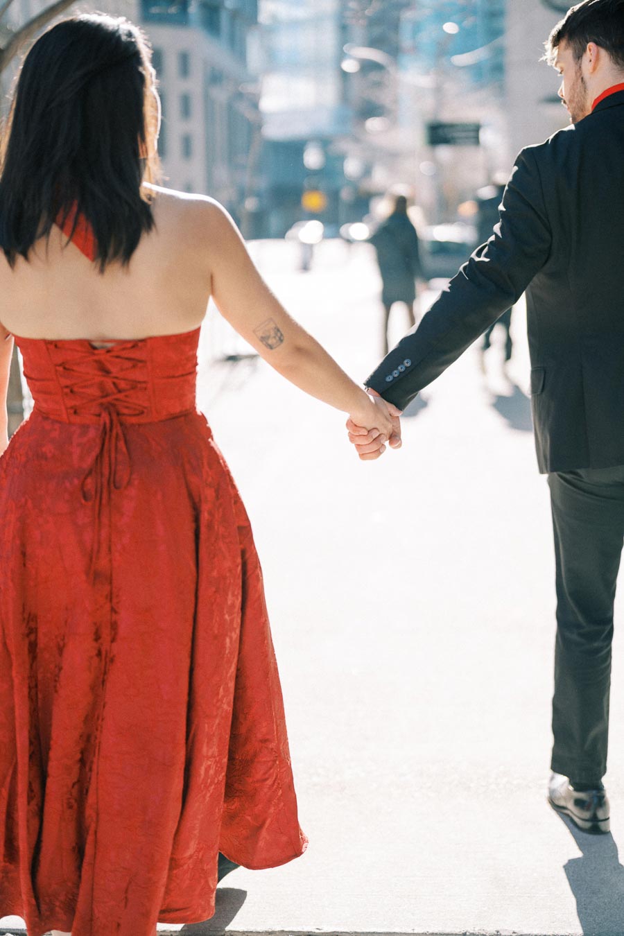 A couple walks hand in hand down a sunlit city street, with the woman in a red dress and the man in a black suit.