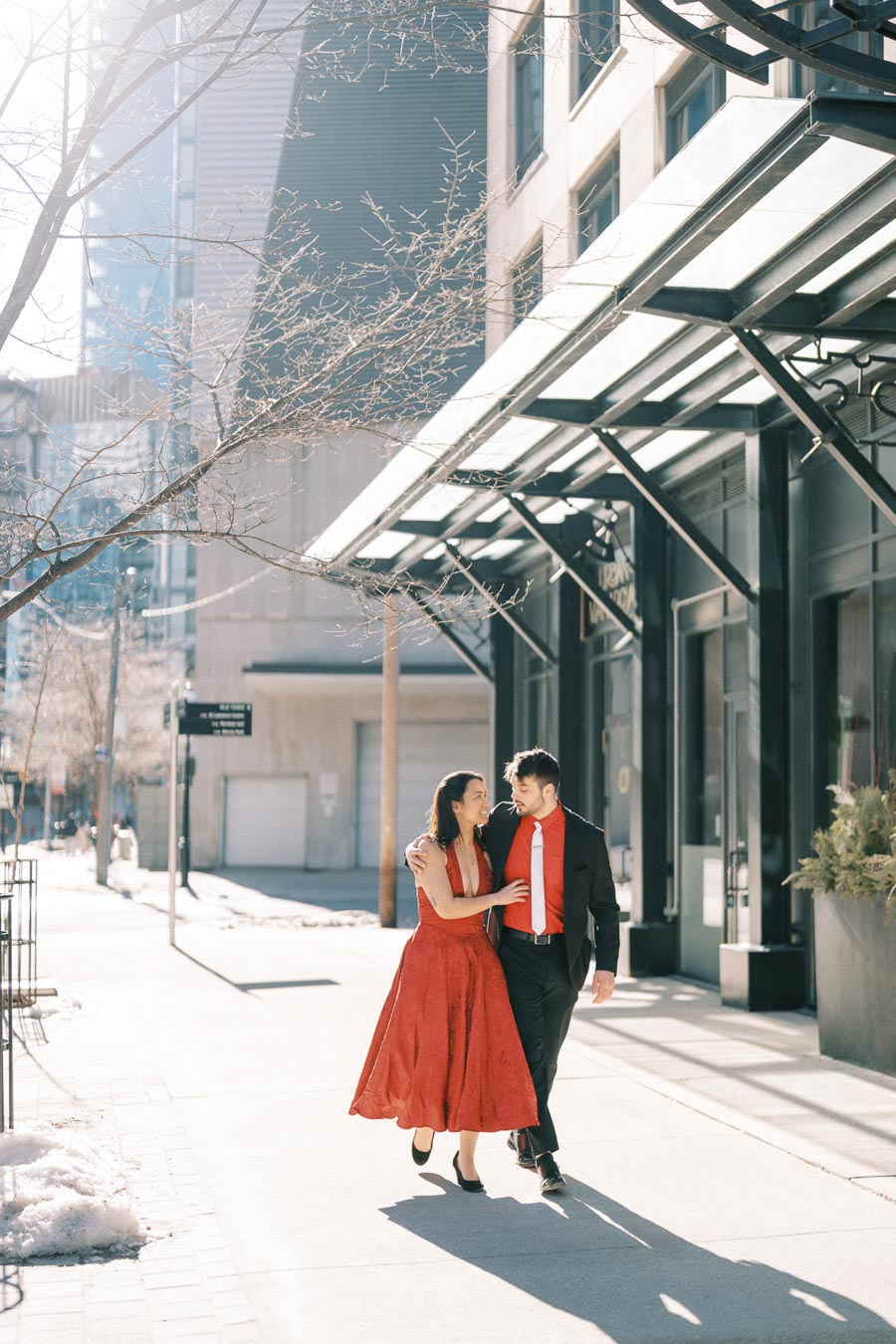 A couple dressed in red walks arm in arm along a sunny city sidewalk, surrounded by modern architecture.