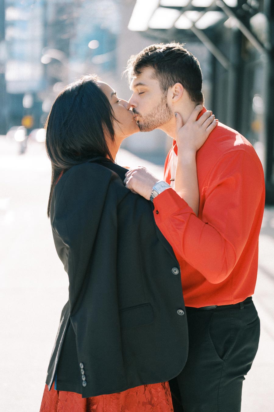 A couple shares a kiss on a sunny city street, dressed in coordinated red and black outfits.