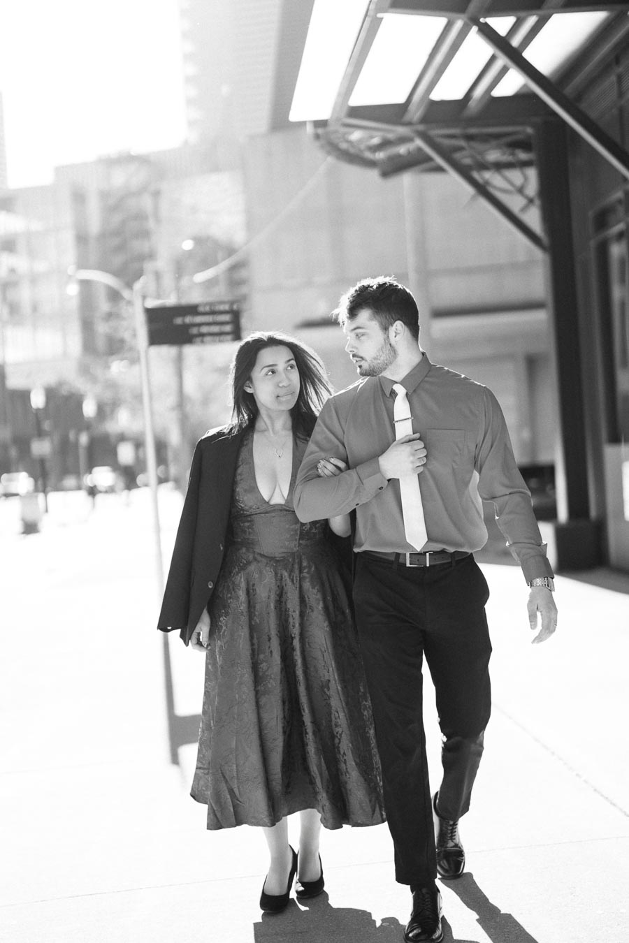 A couple walks arm in arm on a city street, captured in a black and white photograph.