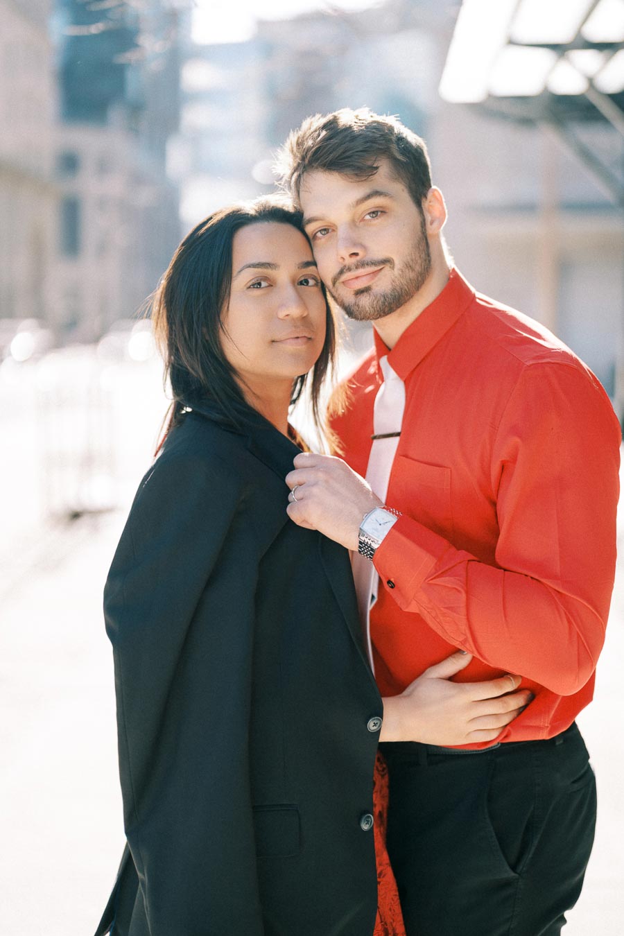 A couple poses affectionately outdoors, with the man in a red shirt and the woman in a black jacket.