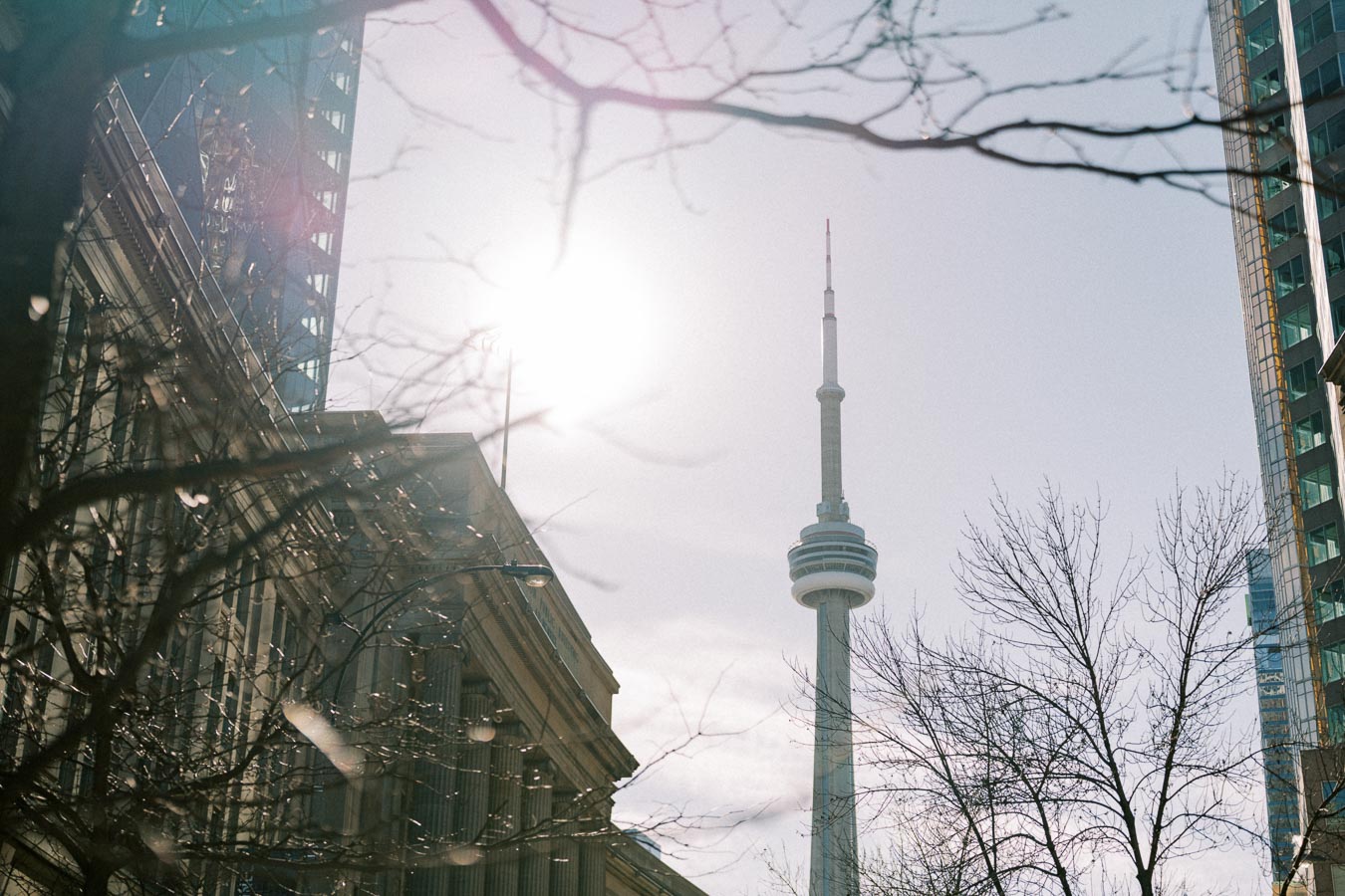 The CN Tower in Toronto is seen through a cityscape of buildings and bare trees, with the sun shining brightly in the sky.