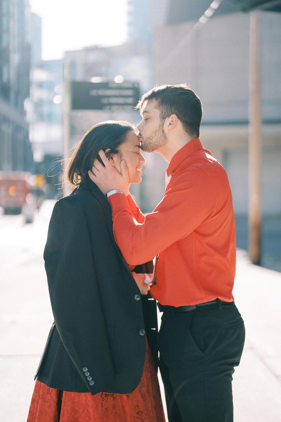 A tender moment captured between a couple on a sunny city street, with the man gently kissing the woman's forehead.