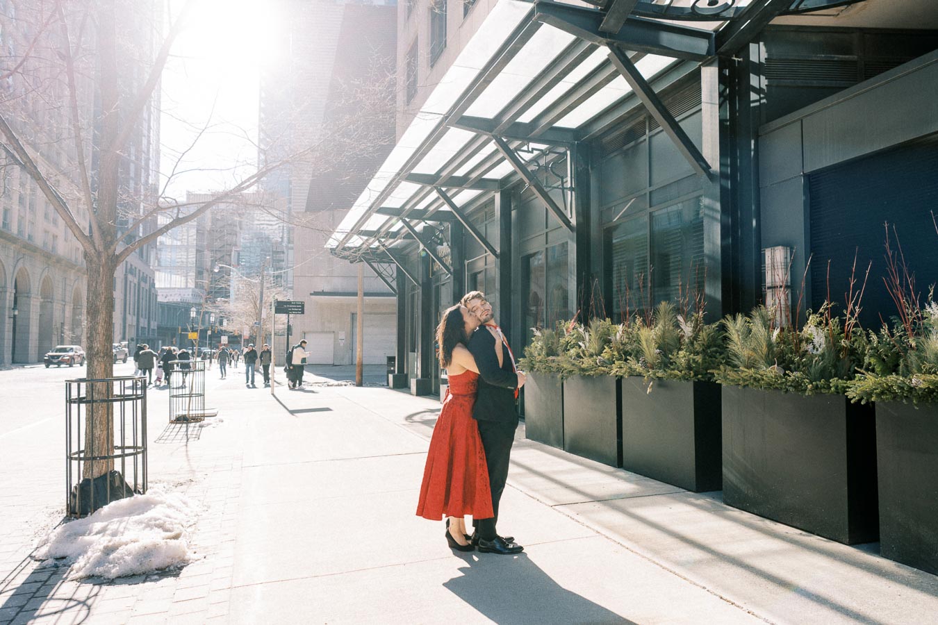 A couple embraces on a sunny city street, surrounded by modern architecture and large planters.