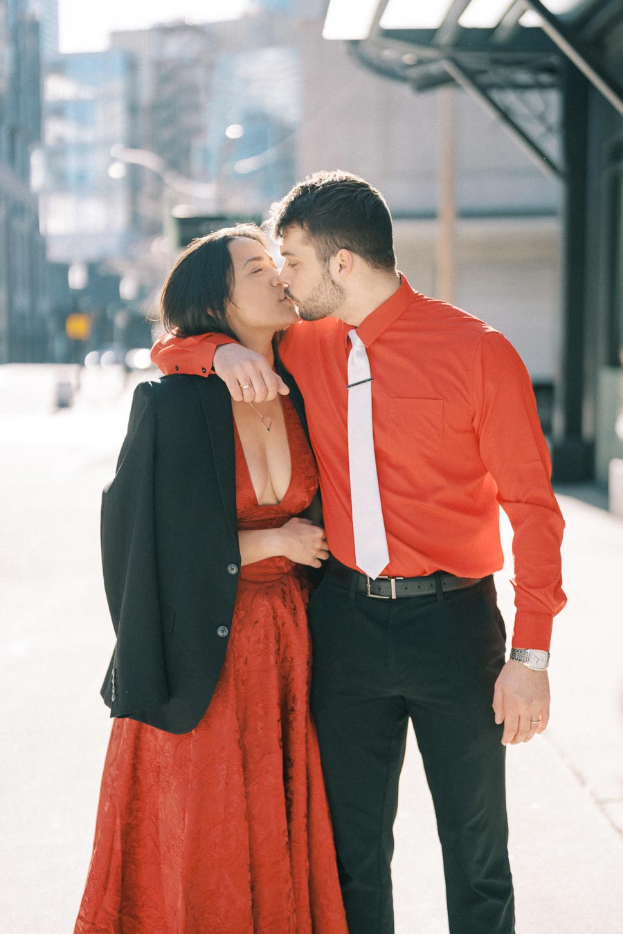 A couple shares a kiss on a sunny city street, both dressed in red attire. The woman wears a red dress with a black jacket, while the man sports a red shirt with a white tie.