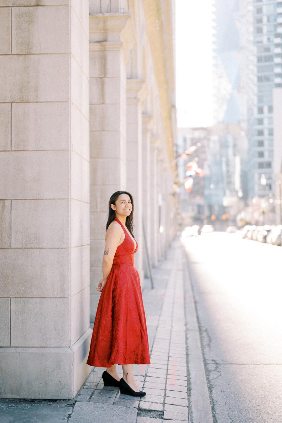 A woman in a red dress stands against a stone wall on a city street, with sunlight illuminating the scene and tall buildings in the background.