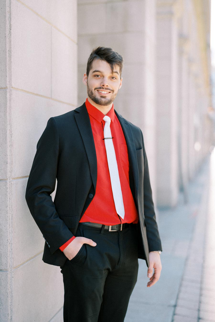A man wearing a black suit with a red shirt and white tie stands confidently against a wall.