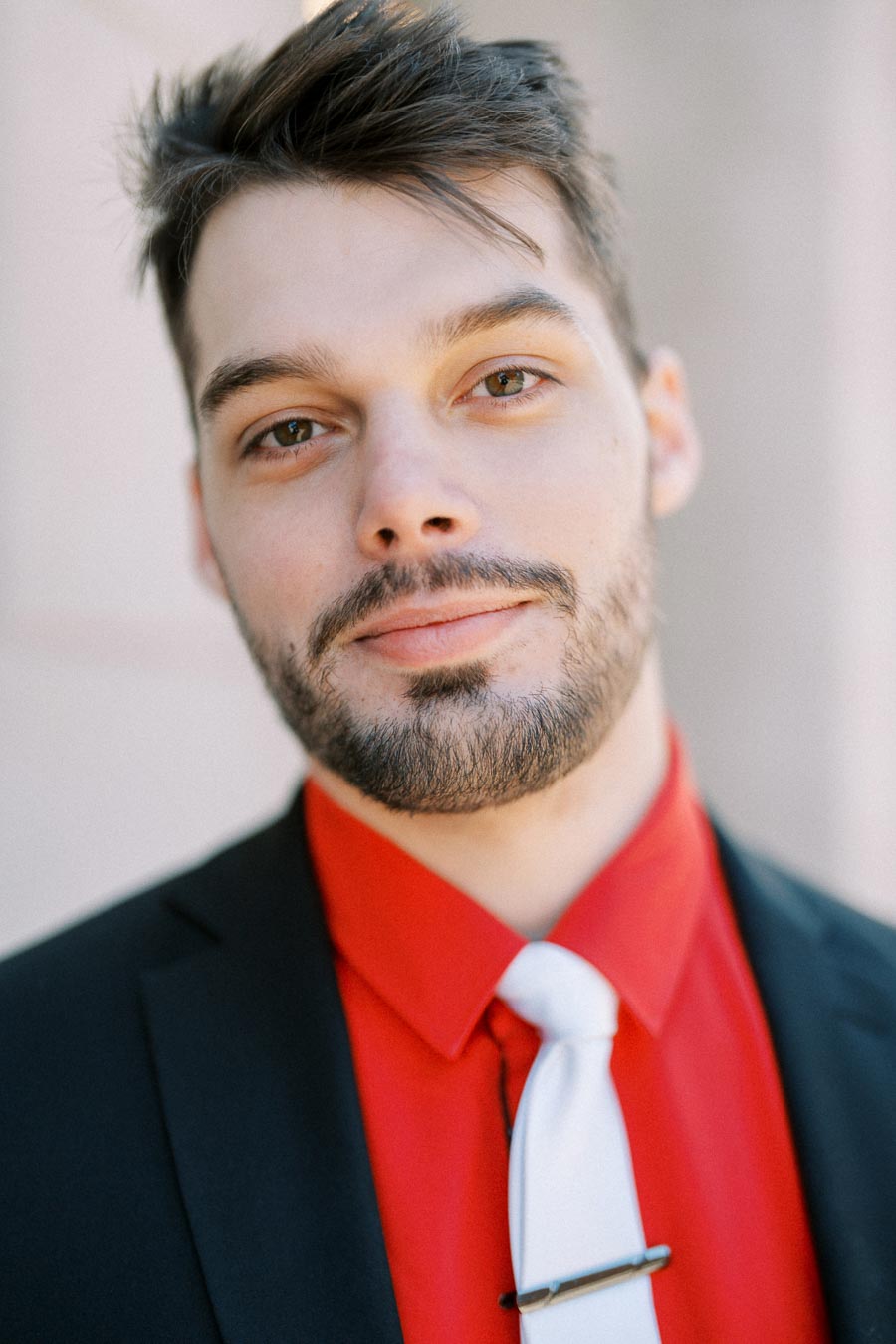 A man dressed in a red shirt, black suit, and white tie, looking directly at the camera.