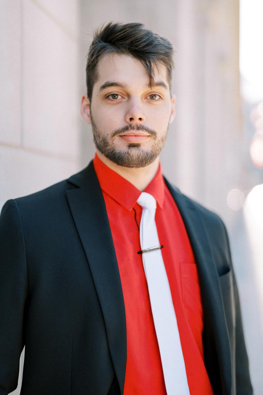 A man wearing a black suit with a red shirt and white tie, posing outdoors.