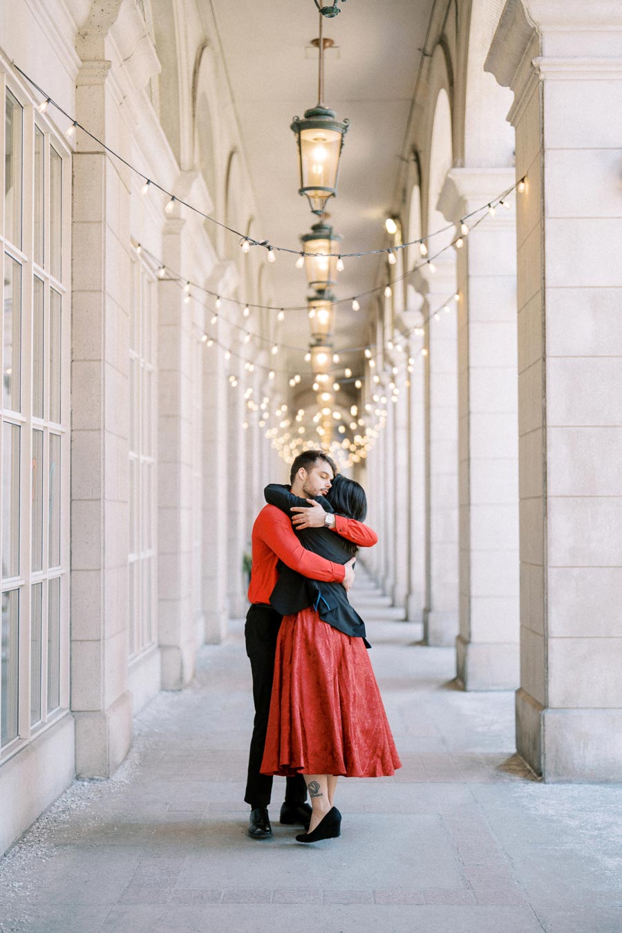 A couple shares an embrace in a beautifully lit corridor adorned with string lights and lanterns, creating a romantic atmosphere.