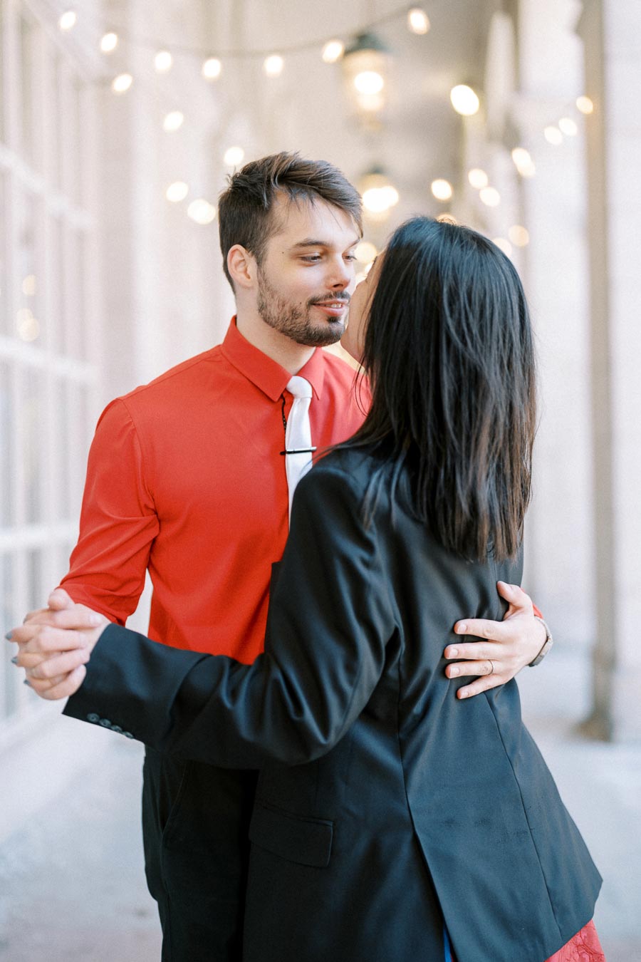 A couple shares a romantic dance under string lights, with the man in a red shirt and the woman in a black jacket.