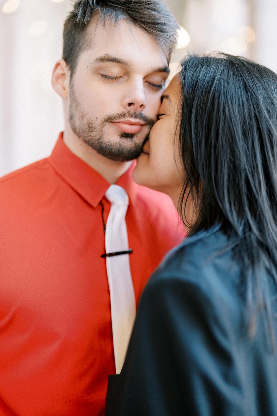 A couple shares an intimate moment, with the man wearing a red shirt and white tie, and the woman in a black outfit.