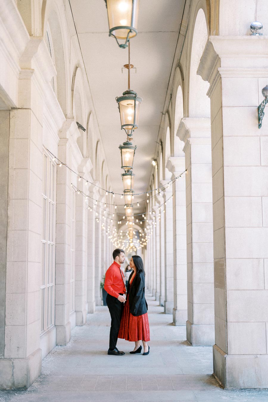 A couple stands in a beautifully lit corridor with arches and hanging lights, creating a romantic atmosphere.