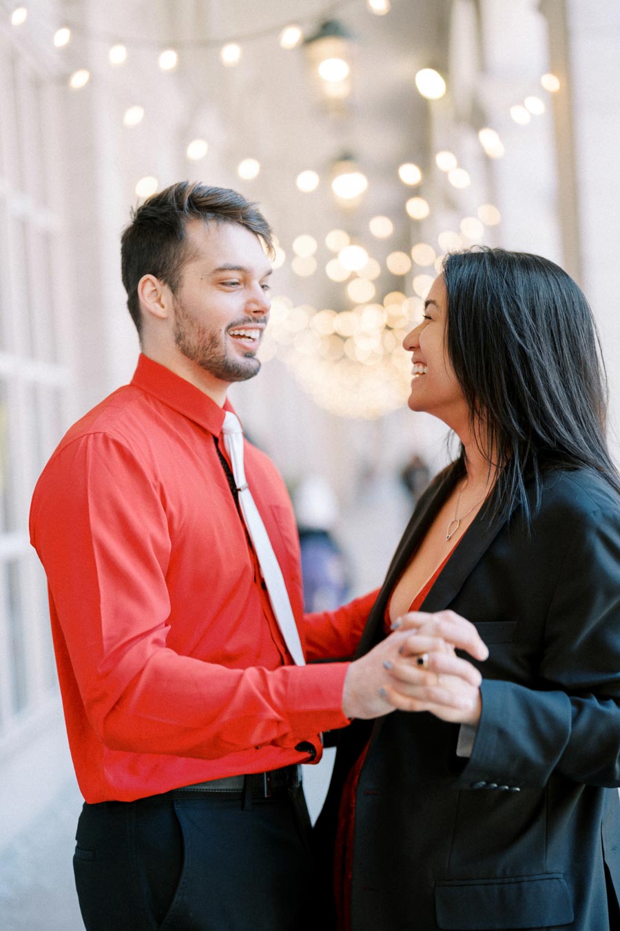A couple shares a joyful moment dancing under string lights, with the man in a red shirt and the woman in a black blazer.