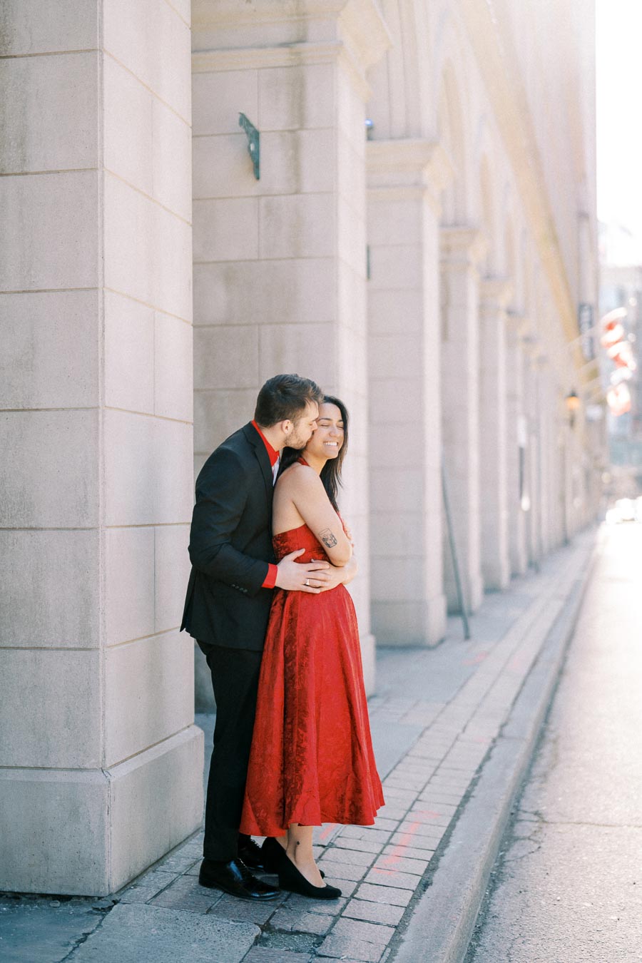 A couple shares a tender moment on a city street, with the man in a black suit and red shirt kissing the woman in a red dress.