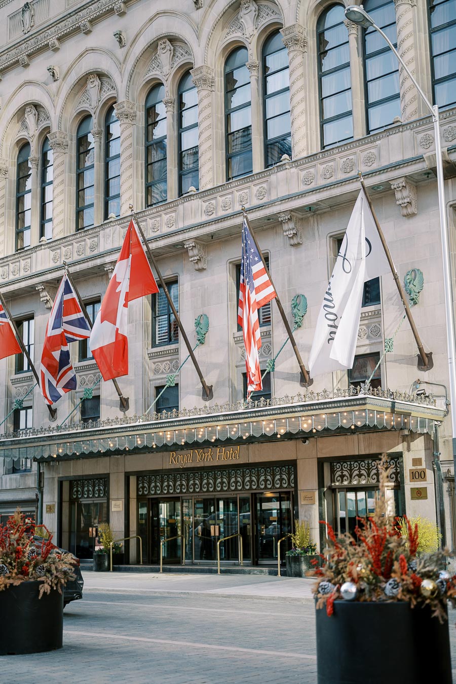 The entrance of the Royal York Hotel features an ornate facade with multiple flags, including those of the United Kingdom, Canada, and the United States. The building's architecture is detailed with arched windows and decorative elements. Planters with seasonal decorations are placed in front of the entrance.