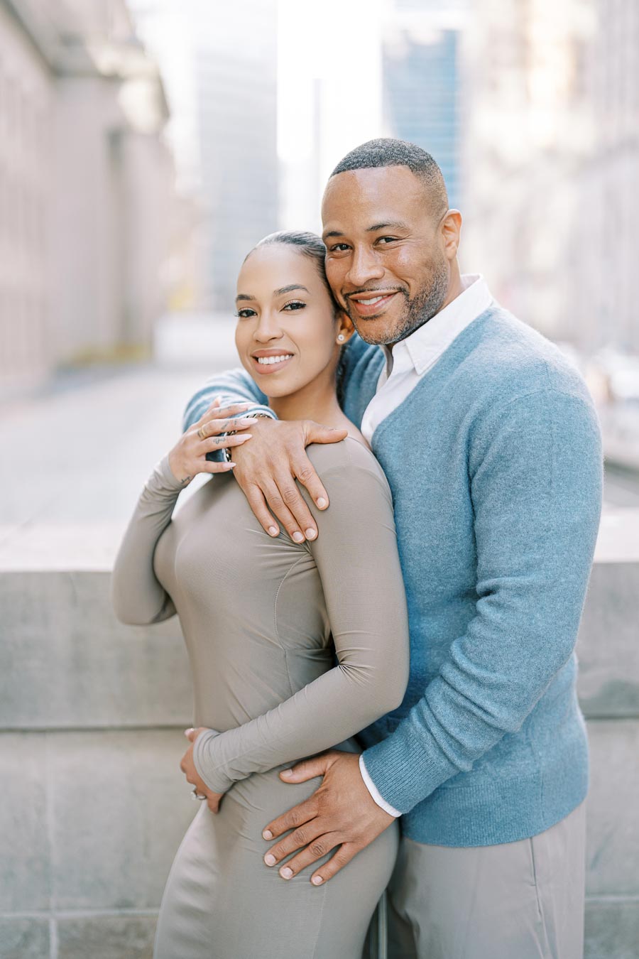A couple embraces and smiles warmly in an urban setting, with tall buildings in the background.
