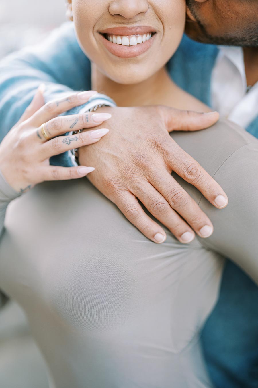 A close-up image of a couple embracing, focusing on their hands and the woman's smile. The woman has tattoos on her fingers and is wearing a ring, while the man has his arm around her shoulder.