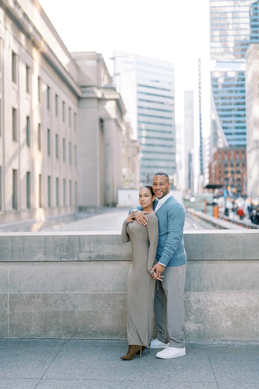 A couple poses affectionately in an urban setting, surrounded by modern skyscrapers.