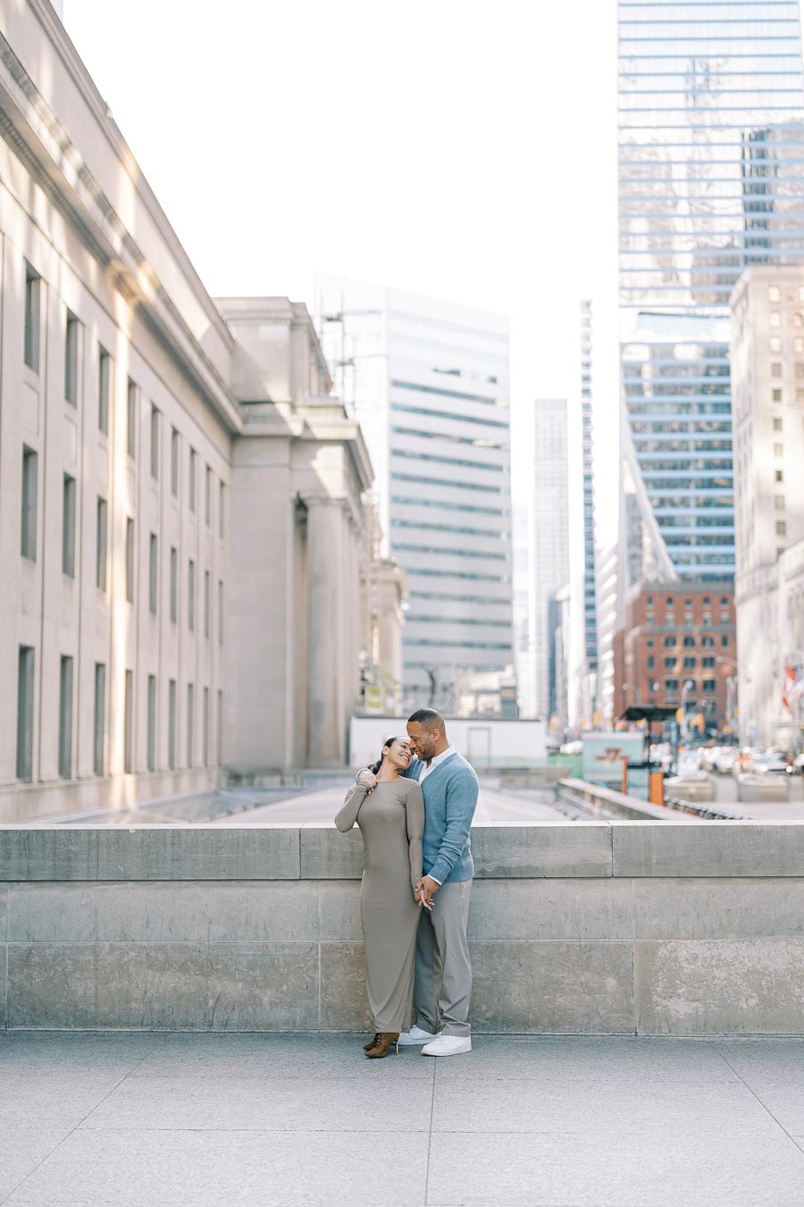 A couple shares a tender moment in an urban setting, surrounded by towering skyscrapers and classic architecture.