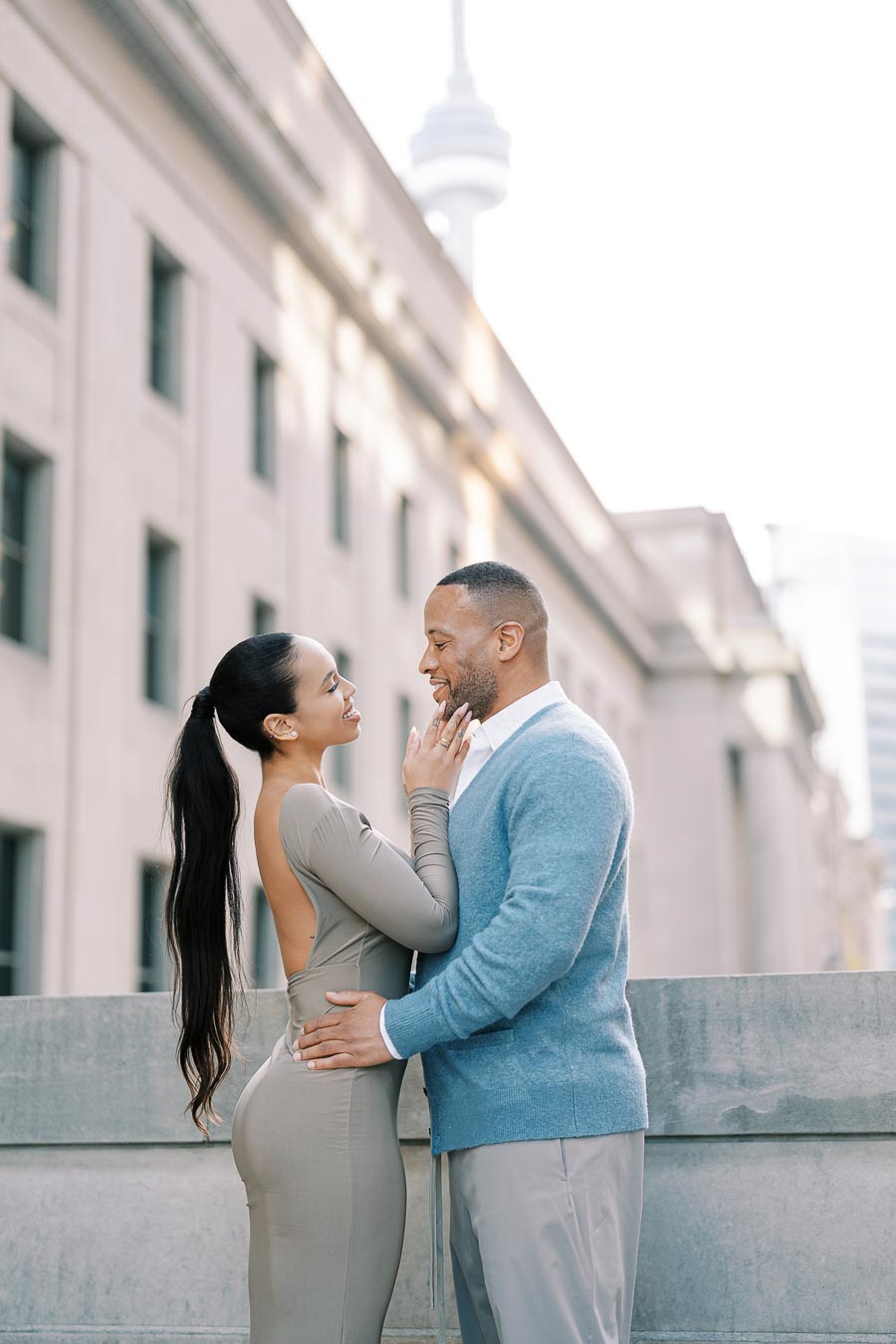 A couple shares a tender moment in front of a building, with the CN Tower visible in the background, suggesting a location in Toronto.