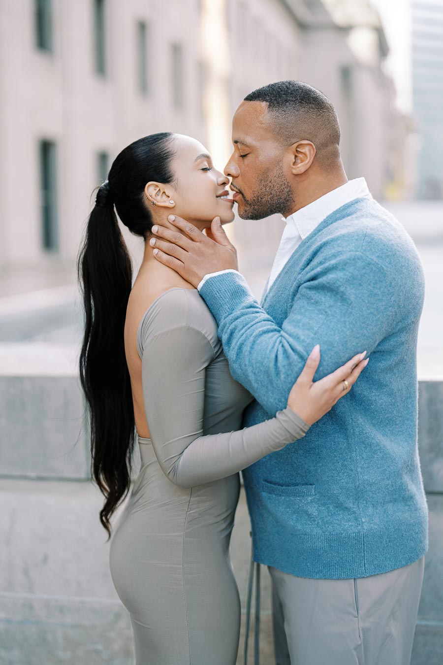 A couple shares a romantic kiss outdoors, with the woman in a form-fitting gray dress and the man in a blue sweater.