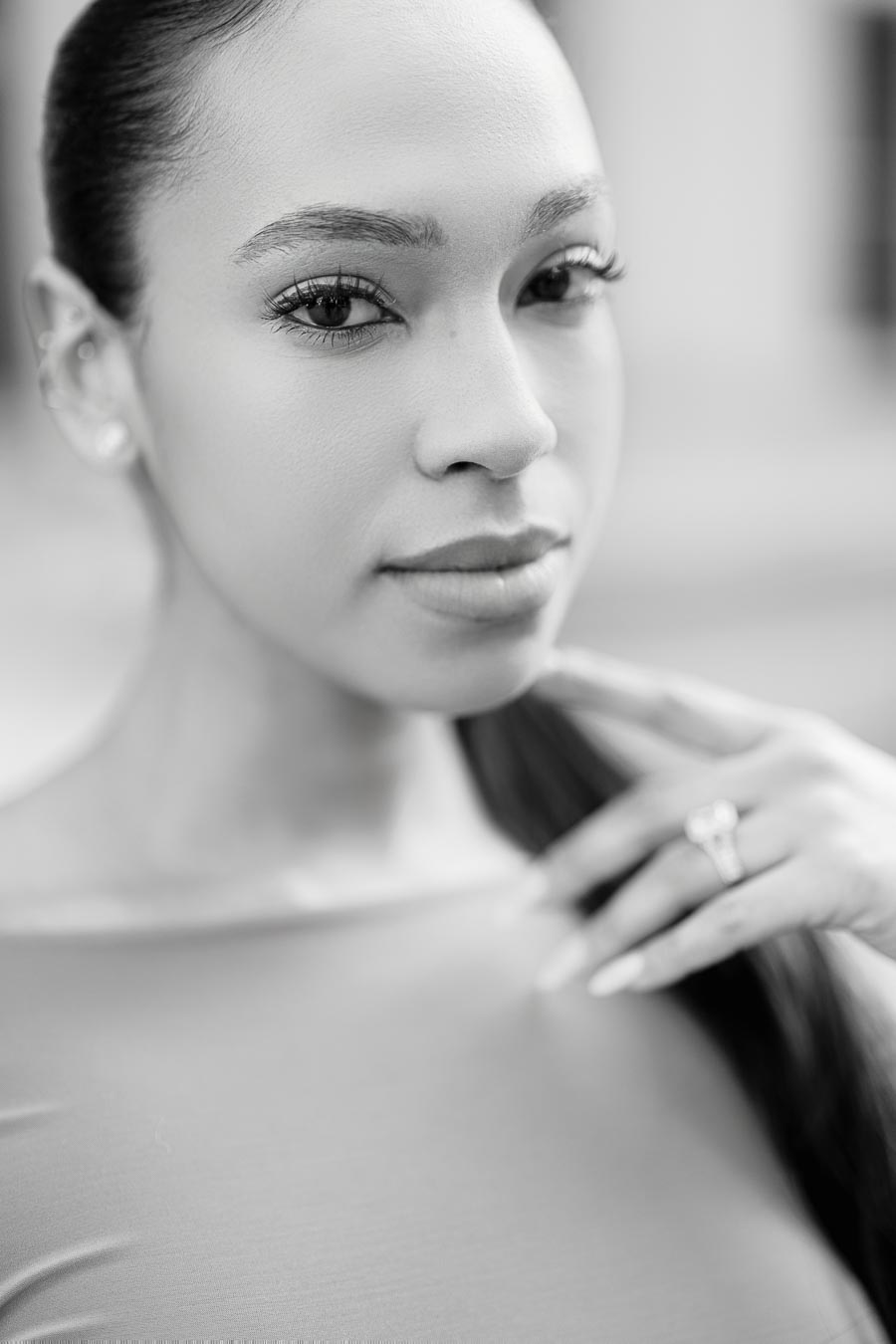 A black and white close-up portrait of a woman with long hair, showcasing her facial features and a ring on her finger.