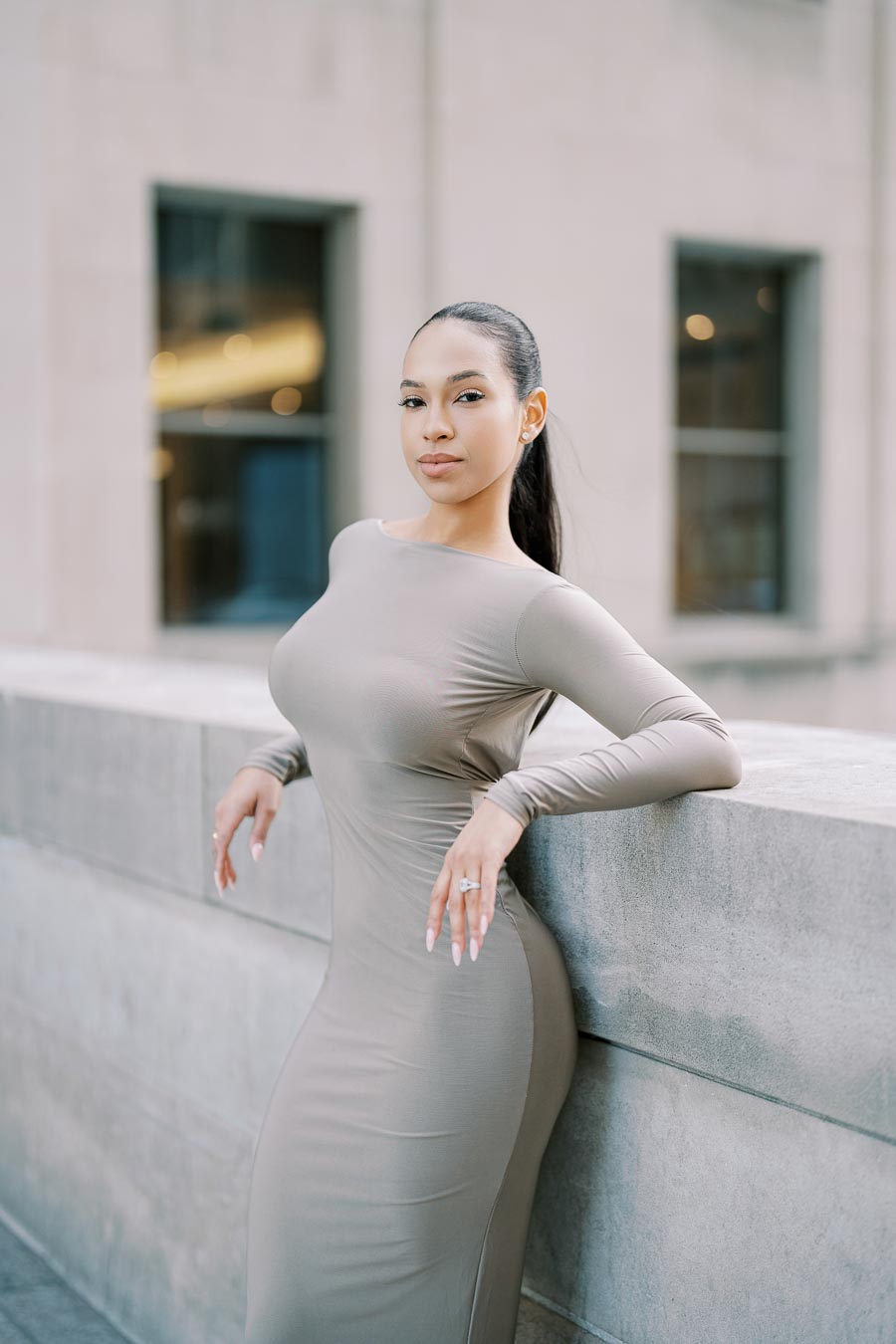 A woman poses confidently in a sleek beige dress, leaning against a concrete wall in an urban environment.