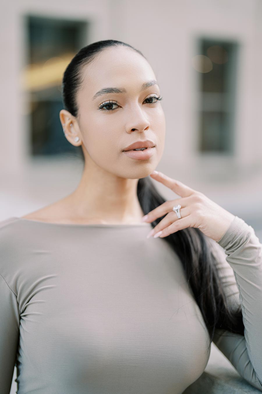 A woman poses elegantly, highlighting her ring and wearing a light gray top.