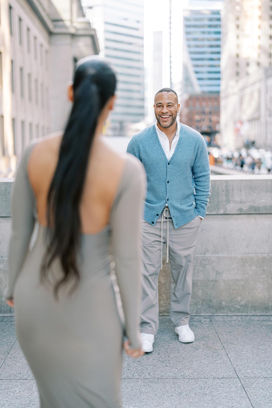 A man in a blue cardigan smiles warmly at a woman in a gray dress on a bustling city street.