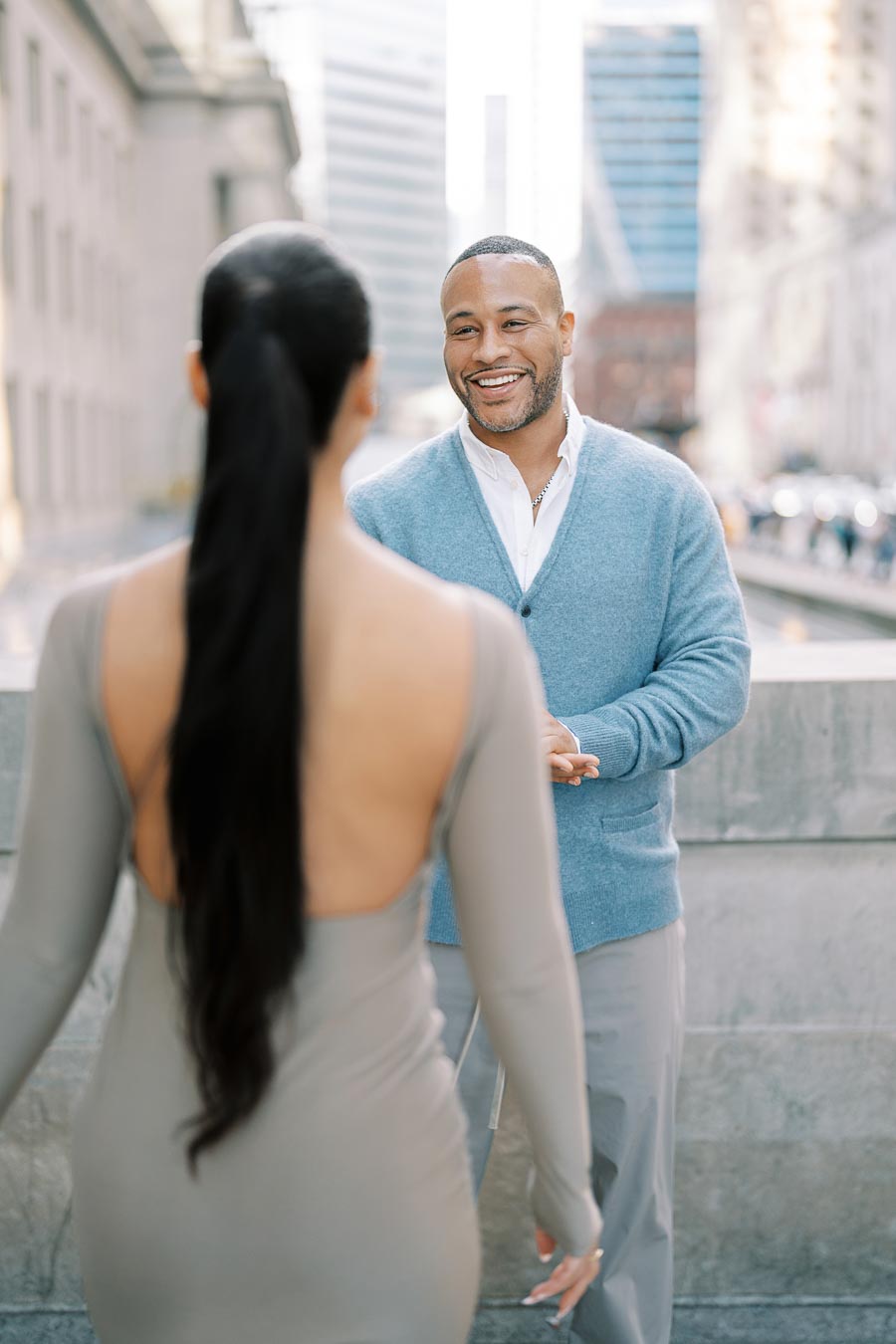 A man in a blue sweater smiles warmly at a woman with long black hair, wearing a backless dress, on a city street.