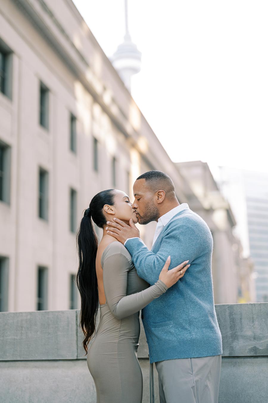 A couple shares a romantic kiss in an urban setting, with the CN Tower visible in the background, suggesting the location is Toronto.