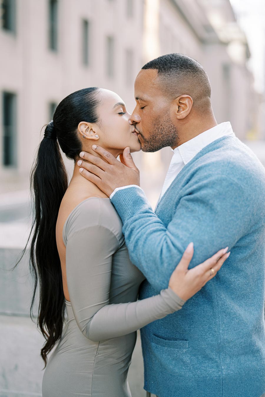 A couple shares a romantic kiss outdoors, with the woman in a gray dress and the man in a blue sweater.