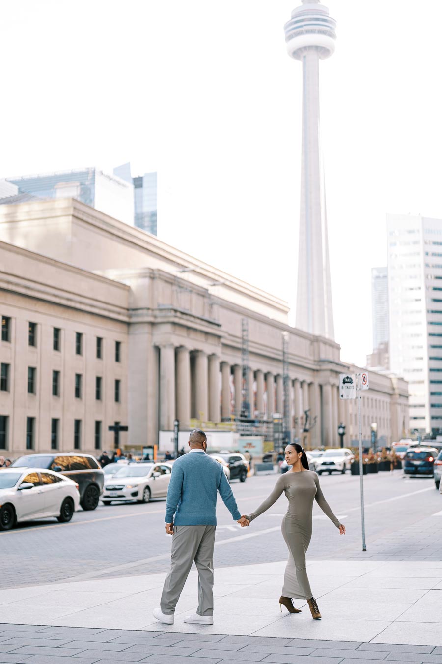 A couple walks hand in hand near Union Station in Toronto, with the CN Tower visible in the background.